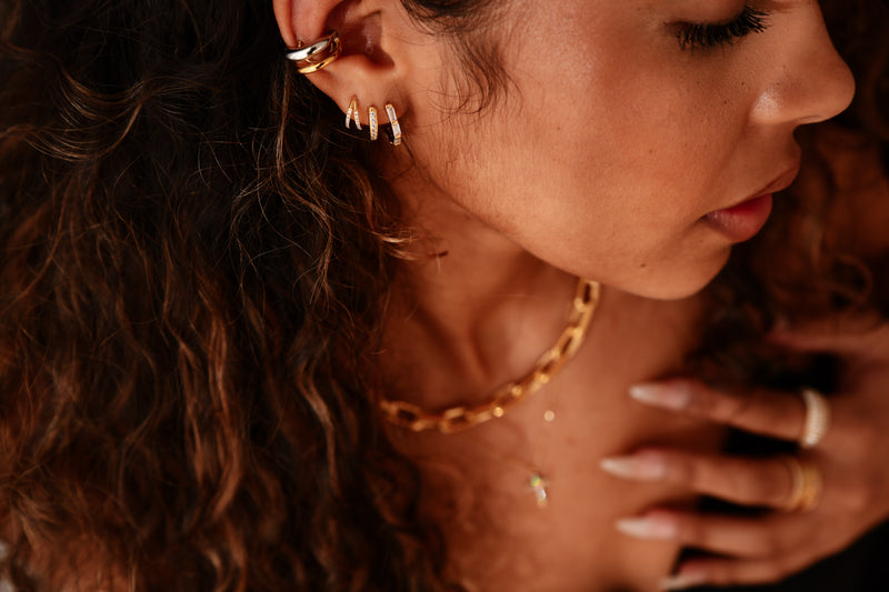 Close-up of a woman's face with gold jewelry on her ear, neck, and hand.