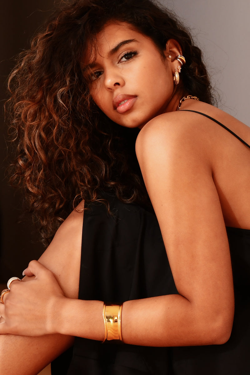 Woman wearing a black top with gold jewelry against a neutral background