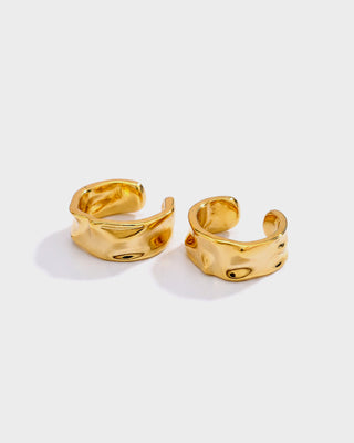 A pair of chunky, organic-shaped gold hoop earrings with a textured, wavy surface, displayed on a clean light gray background.