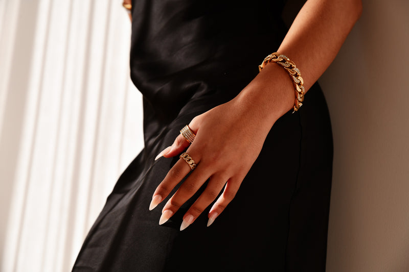 Hand wearing a gold bracelet and ring on a black garment with a neutral background